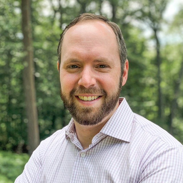 Andrew White in a pinstripe shirt standing in the woods 