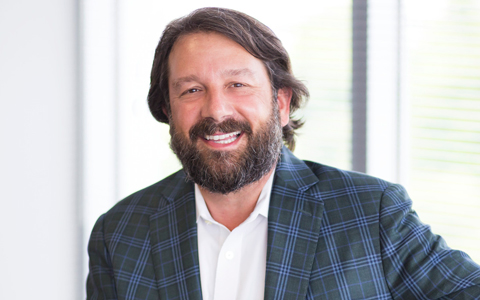 Kris Dramby, wearing a white shirt and plaid blazer, smiles in the office.