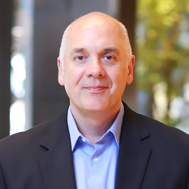 Joe wearing a navy blazer and blue shirt in front of a blurred background.