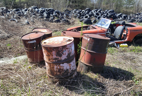 Grassy lot with rusted barrels, scrapped car, and large pile of tires.