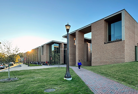 A woman in a blue shirt walks along a path in front of a brick and glass college performing arts center.