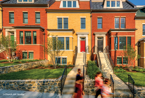 A group of people walking past stairs leading to a row of multi-story red and yellow brick townhomes.