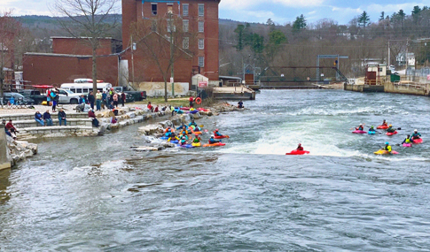 Numerous kayakers in multi-colored kayaks paddle in river rapids near a spectator viewing area.