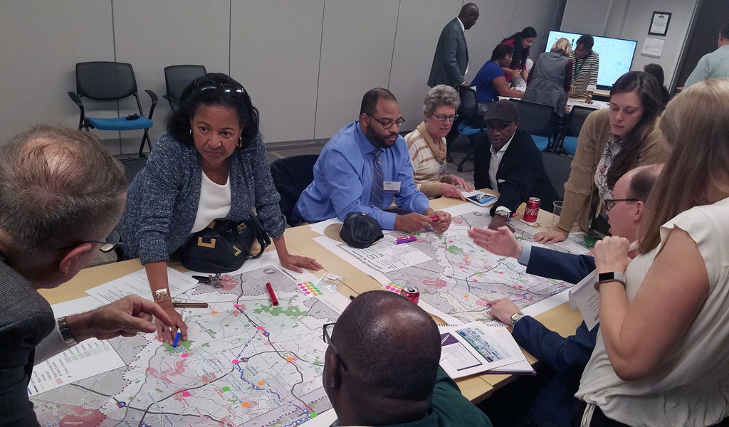 A diverse group of people sit and stand around a table looking at two maps of transit routes. 