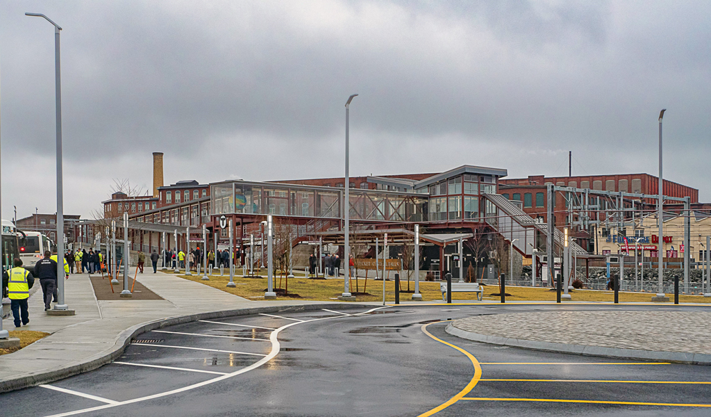 People gather and walk around the exterior of brick train station with glass walkways and windows.