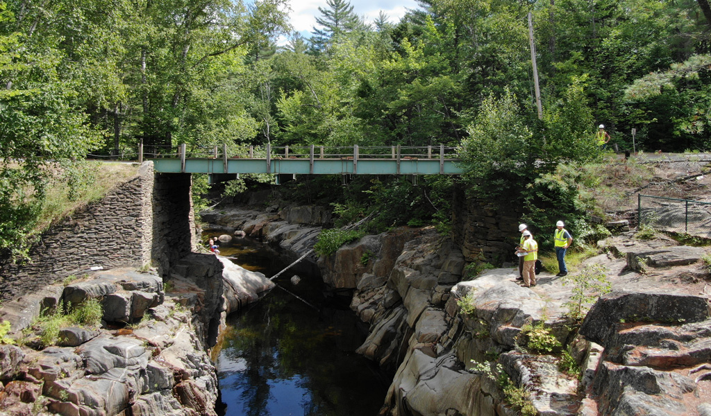 Three men stand on a small cliffside under a bridge above a large stream.