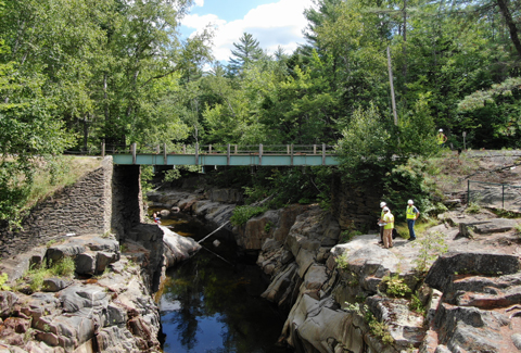 Three men stand on a small cliffside under a bridge above a large stream.