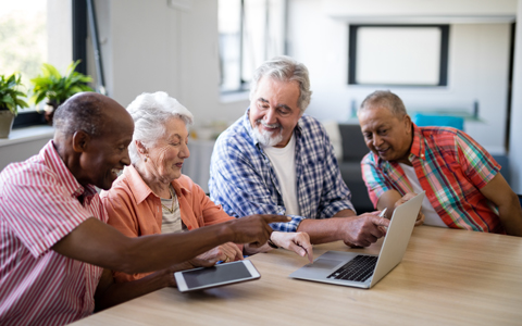 A group of elderly people are sitting at a table looking at a laptop computer and tablet together.