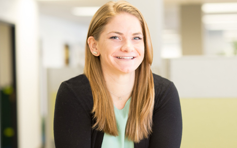 Headshot of Kelly Siry, a woman wearing a blue top and black cardigan smiling while standing in an office. 