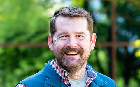 Headshot of Ryan Scott, a man wearing a plaid shirt and a vest smiling with trees in the background. 