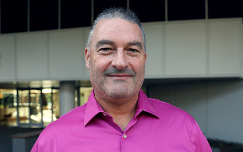 Todd stands  in front of a building wearing a pink shirt