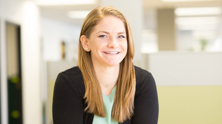 Headshot of Kelly Siry, a woman wearing a blue top and black cardigan smiling while standing in an office. 