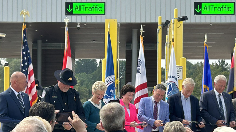 A group of officials cutting a ribbon in front of flags.   