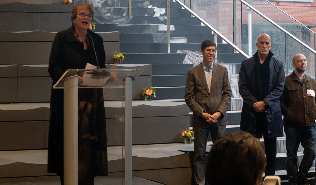 A woman stands at a podium in front of a large staircase, delivering a speech with colleagues standing to her right. 