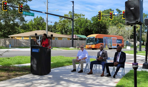 Three people are seated by a standing speaker at a podium on a path.