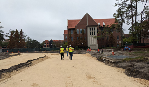 Three people wearing construction hard harts and yellow vests walking over a construction site covered with sand.