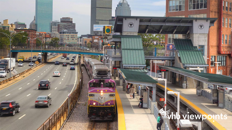 Commuters wait on a platform as a train pulls into the station, with the City of Boston in the background.